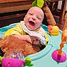 baby, infant, child, laughing, smiling, face, portrait, activity_walker, baby_toy, colorful_toys, plush_toy, indoor, playtime, seated, yellow_clothing, bib, wooden_table, furniture, happy, photograph