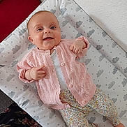Charley is registered to the contest to win money with this photo: baby, smiling, infant, pink_sweater, floral_pants, cushioned_pad, cloud_pattern, star_pattern, happy, lying_down, cute, child, person, indoor, soft_fabric, cozy, small_hands, small_feet, wall, red_cushion
