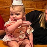 baby, infant, child, birthday, party_hat, pink_clothing, smiling, sitting, portrait, family, adult, faces, wooden_background, indoors, celebration, cake_absent, baby_dress, lace, holding, happy