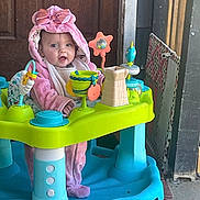 Sadie joined the competition — help win amazing prizes! baby, child, infant, pink_hood, bow, smiling, sitting, activity_center, baby_toys, colorful, toy_fortress, portrait, indoors, doorway, wooden_door, playtime, happy, cute, bright_colors, floor_tiles