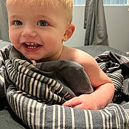 Jaxson is registered to the contest to win money with this photo: toddler, child, smiling, blanket, striped_blanket, indoor, bed, curtains, window, blonde_hair, blue_eyes, happy, cute, person, skin, hand, portrait, cozy, soft_light, young