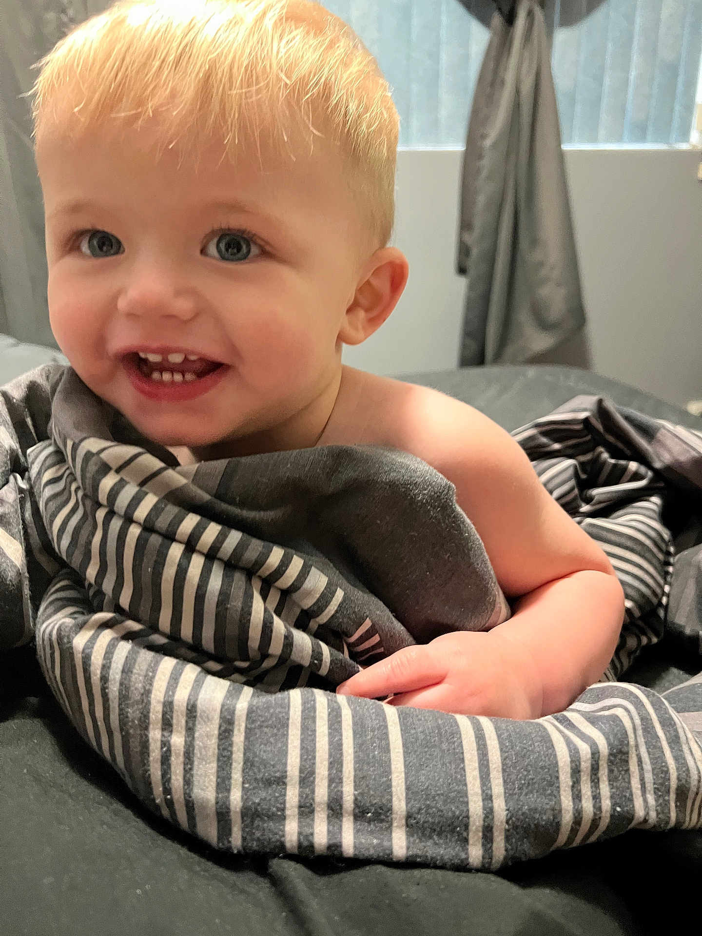 Jaxson is registered to the contest to win money with this photo: toddler, child, smiling, blanket, striped_blanket, indoor, bed, curtains, window, blonde_hair, blue_eyes, happy, cute, person, skin, hand, portrait, cozy, soft_light, young