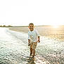 child, boy, water, beach, sunlight, smile, sand, shore, nature, happy, summer, outdoor, casual_clothing, shorts, white_shirt, reflection, waves, sunset, playful, portrait