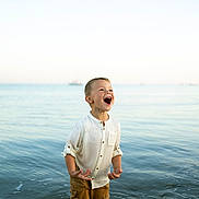 Côme participe au concours pour gagner de l'argent avec cette photo : child, boy, water, ocean, shore, laughing, happy, shorts, shirt, summer, outdoor, nature, sea, waves, sky, sunlight, vacation, playful, standing, daytime