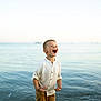 child, boy, water, ocean, shore, laughing, happy, shorts, shirt, summer, outdoor, nature, sea, waves, sky, sunlight, vacation, playful, standing, daytime