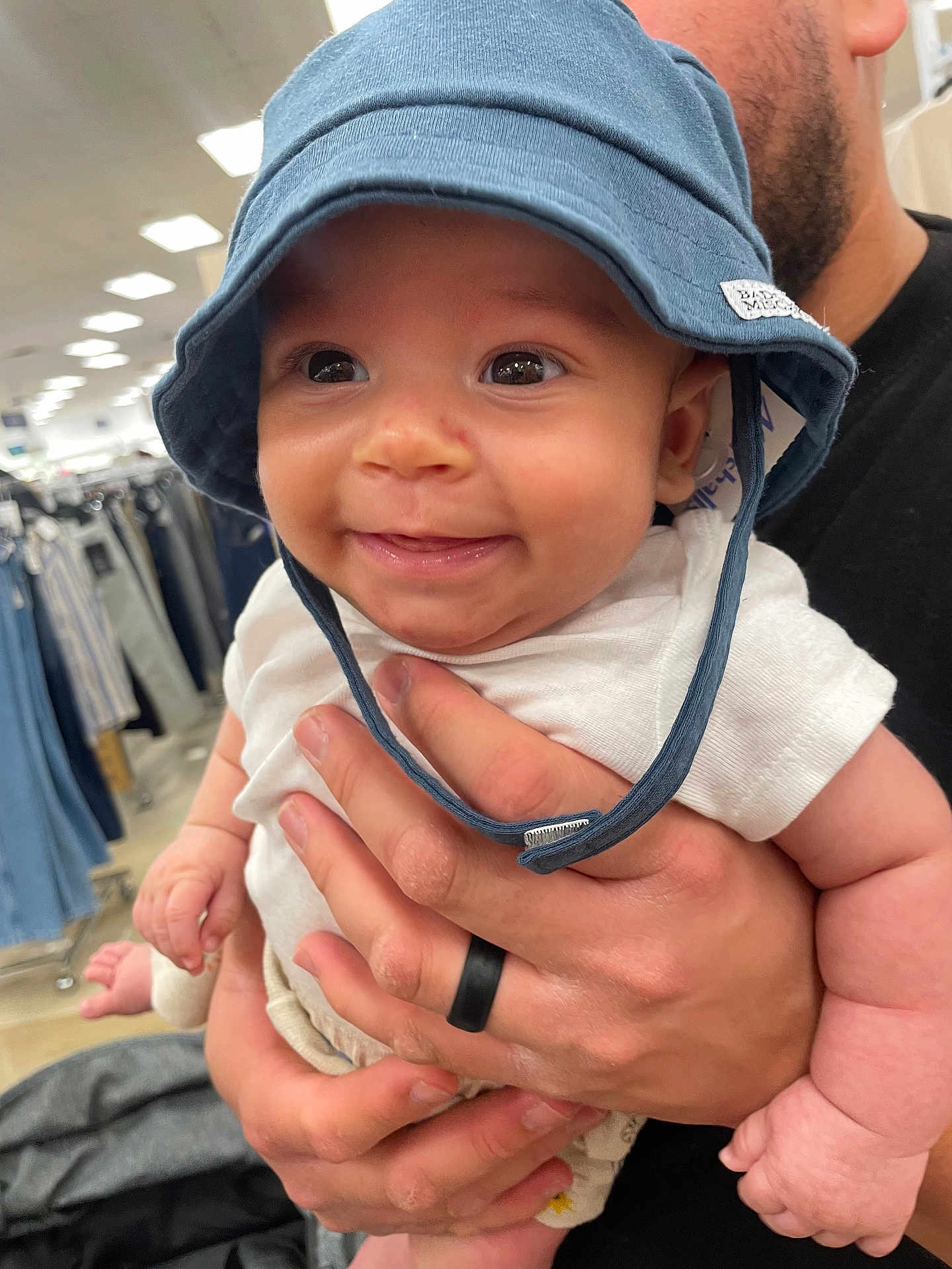 Jalen is registered to the contest to win money with this photo: baby, child, hat, blue_hat, smile, happy, person, hand, adult, clothing_store, shirt, white_shirt, ring, black_ring, holding, retail, shopping, aisle, indoor, face