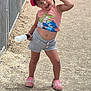child, girl, cap, tank_top, shorts, pink_crocs, water_bottle, outdoor, dirt_path, playful, pose, smile, face, person, summer_clothing, daylight, fence, cute, happy, casual