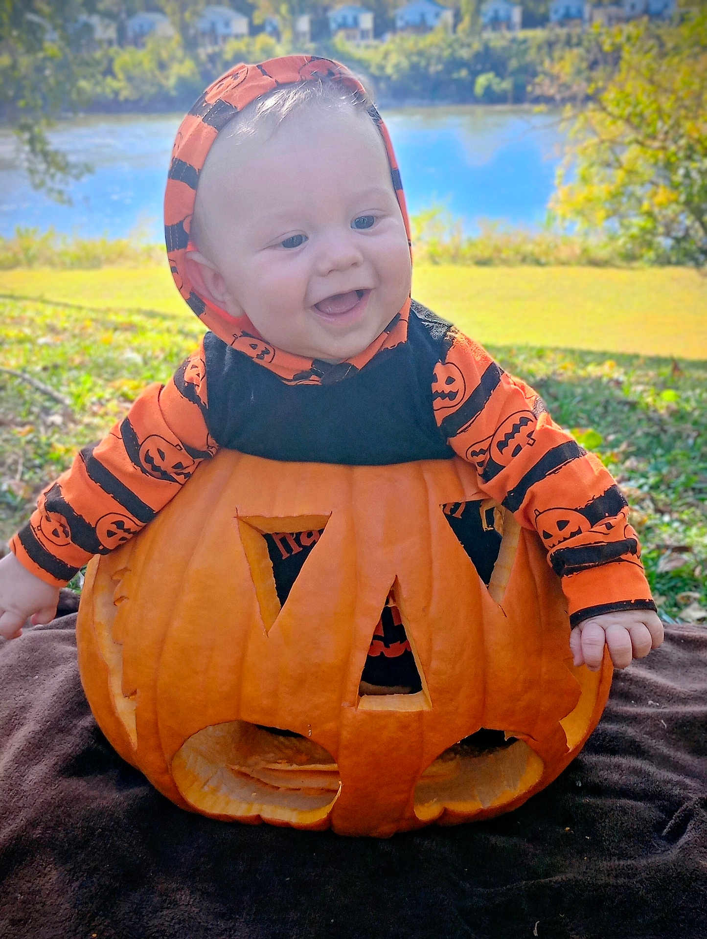Leland is registered to the contest to win money with this photo: baby, pumpkin, jack_o_lantern, smiling, hoodie, orange, black, outdoor, grass, lake, trees, fall, seasonal, holiday, cute, child, face, happy, nature, costume