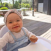 Myla participe au concours pour gagner de l'argent avec cette photo : baby, child, smile, high_chair, headband, denim_overalls, bread, outdoor, garden, patio, table, chair, sunlight, happy, person, infant, nature, daylight, cute, food