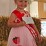 child, girl, dress, strawberry, tiara, sash, smile, hay_bale, envelope, shoes, socks, indoor, fair, award, prince, red, pink, happy, event, standing