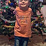 child, christmas_tree, lights, ornaments, orange_shirt, jeans, bows, smile, holiday, indoor, carpet, decorations, happy, person, festive, celebration, tree_stand, home, tinsel, young