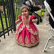 Kheylani Sophia is registered to the contest to win money with this photo: toddler, child, dress, pink_dress, tiara, outdoor, stroller, fence, concrete_floor, smiling, happy, person, baby, royal_costume, footwear, grass, metal_fence, chair, daylight, portrait