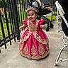 toddler, child, dress, pink_dress, tiara, outdoor, stroller, fence, concrete_floor, smiling, happy, person, baby, royal_costume, footwear, grass, metal_fence, chair, daylight, portrait
