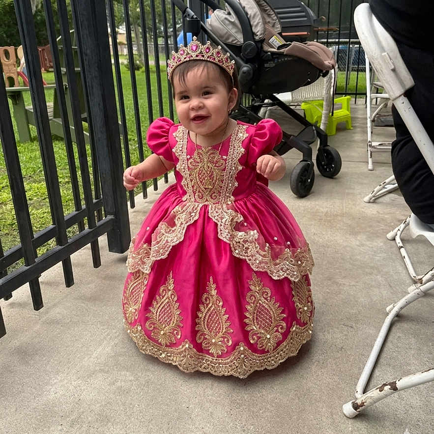 Kheylani Sophia is registered to the contest to win money with this photo: toddler, child, dress, pink_dress, tiara, outdoor, stroller, fence, concrete_floor, smiling, happy, person, baby, royal_costume, footwear, grass, metal_fence, chair, daylight, portrait