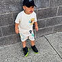 black_shoes, boy, brick_wall, cap, casual_clothing, child, concrete, curious_expression, daylight, headwear, mickey_mouse, nike, outdoor, shorts, sneakers, standing, street_style, tshirt, urban, young_child