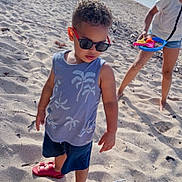 Pedro is registered to the contest to win money with this photo: background, beach, child, coast, ocean, outdoor, person, red_sandals, sand, shadow, shorts, sky, summer, sunglasses, sunlight, tank_top, toddler, toys, vacation, water