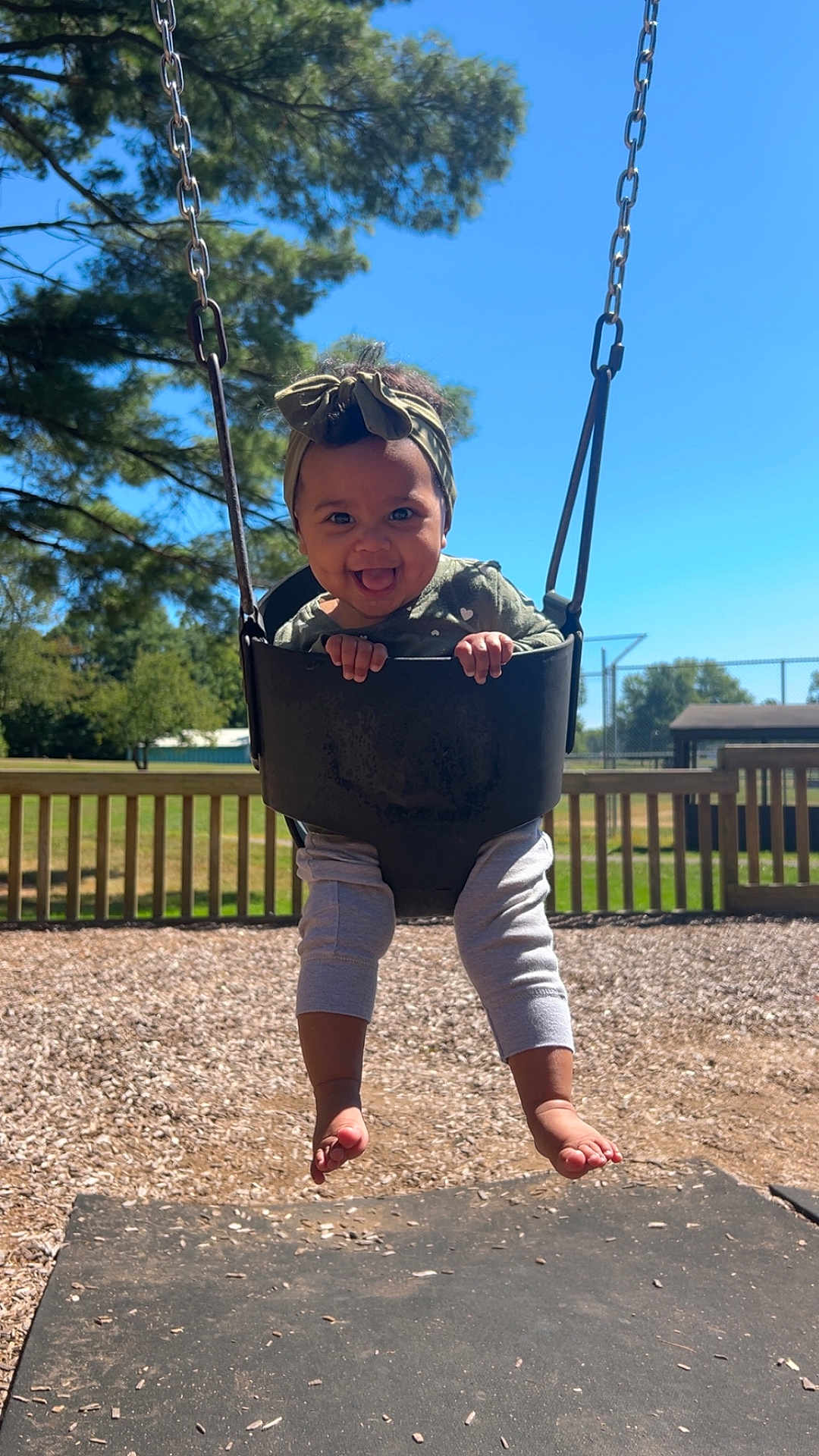 Anaya is registered to the contest to win money with this photo: child, toddler, swing, playground, outdoor, smiling, happy, headband, tree, sky, sunlight, grass, fence, barefoot, clothing, person, fun, play, woodchips, daytime