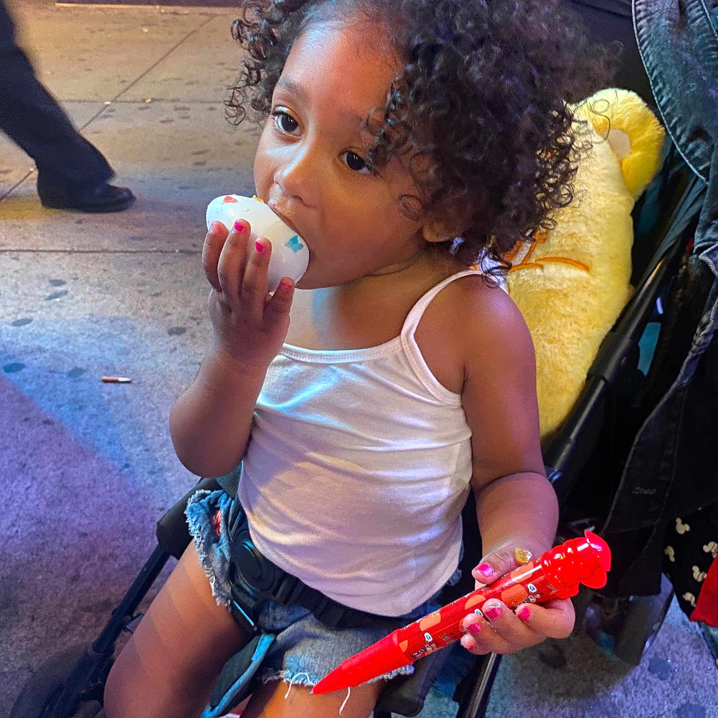 Amiyah is registered to the contest to win money with this photo: toddler, child, curly_hair, stroller, toy, red_pen, plush_toy, sidewalk, urban, night, casual_clothing, white_tank_top, denim_shorts, nail_polish, food, snack, outdoor, person, cute, playful