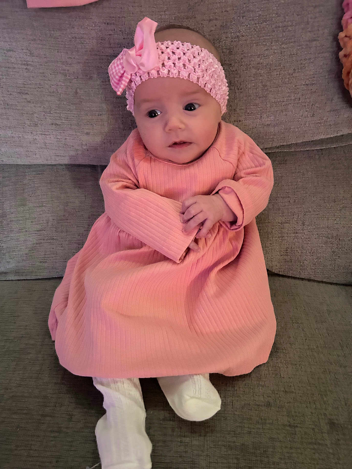 Chelsea joined the competition — help win amazing prizes! baby, infant, pink_dress, headband, couch, gray_couch, white_sock, clothing, hands, face, person, seated, indoor, cute, young_child, portrait, soft_texture, looking_away, curious, small