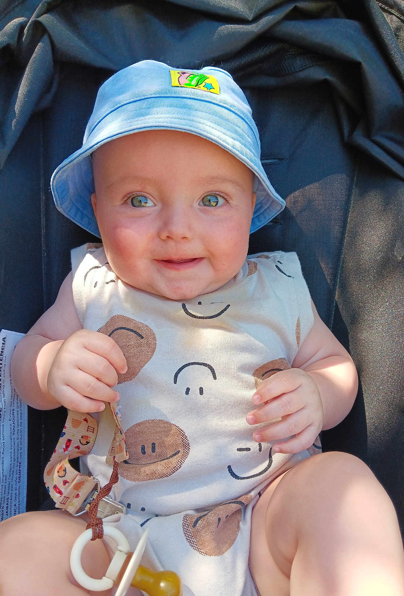 Bentley is registered to the contest to win money with this photo: baby, child, smiling, blue_hat, onesie, pacifier, stroller, infant, cute, happy, outdoor, sunlight, hands, face, eyes, cheeks, clothing, sitting, toy, closeup