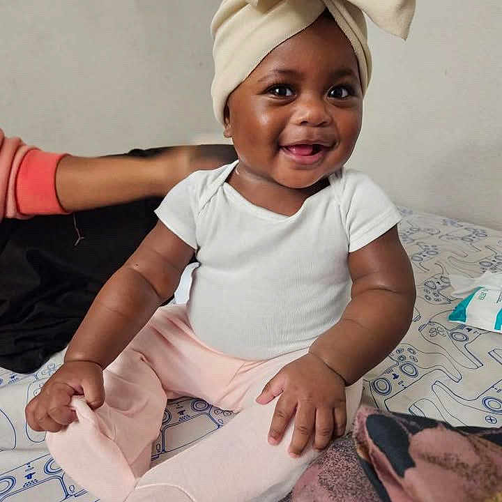Imoni joined the competition — help win amazing prizes! baby, bed, bow, child, clothing, cute, foot, hand, happy, headwear, indoor, infant, patterned_bedspread, person, pink_pants, rainbow_patch, seated, skin, smiling, white_onesie