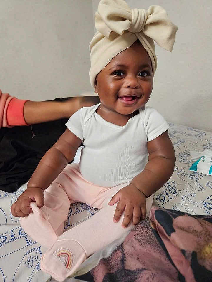 Imoni joined the competition — help win amazing prizes! baby, child, smiling, bow, headwear, white_onesie, pink_pants, rainbow_patch, bed, patterned_bedspread, indoor, person, cute, happy, infant, skin, hand, foot, seated, clothing