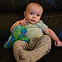 baby, child, plush_toy, dinosaur_toy, sofa, leather, clothing, pants, tshirt, sitting, indoor, person, cute, infant, young_child, face, expression, hand, foot, cozy