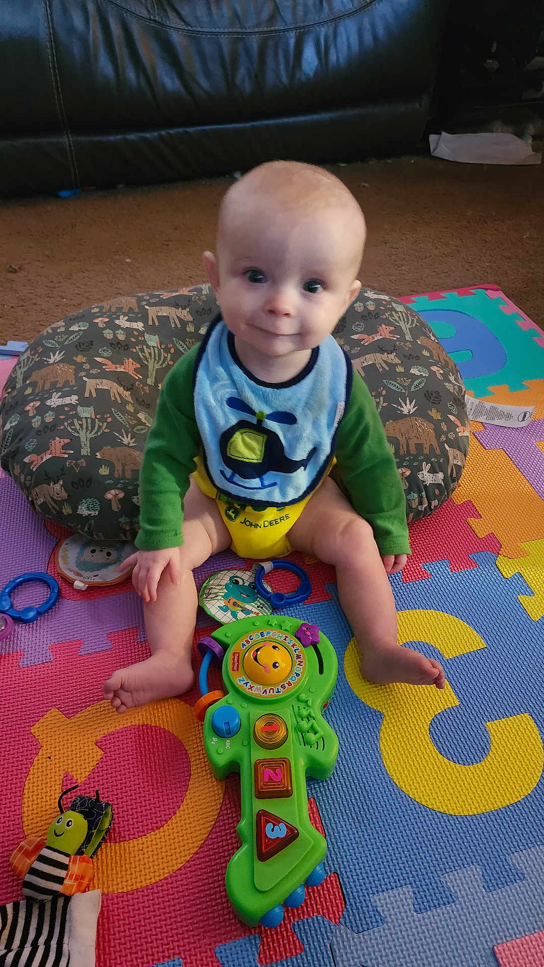 Corsen joined the competition — help win amazing prizes! baby, child, toy, guitar_toy, bib, pillow, foam_mat, carpet, smiling, sitting, indoors, colorful, green_clothing, yellow_clothing, blue_bib, playtime, happy, infant, face, person