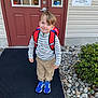 child, boy, backpack, blue_shoes, standing, door, red_backpack, smiling, outdoor, rocks, plants, sign, clothing, casual_wear, happy, sidewalk, building, window, door_handle, long_sleeve_shirt