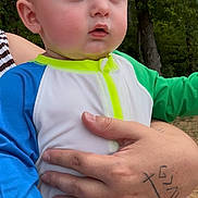 Myles is registered to the contest to win money with this photo: baby, child, infant, person, hand, tattoo, outdoor, nature, trees, clothing, blue_sleeve, green_sleeve, white_outfit, expression, portrait, skin, face, holding, adult, background