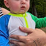 baby, child, infant, person, hand, tattoo, outdoor, nature, trees, clothing, blue_sleeve, green_sleeve, white_outfit, expression, portrait, skin, face, holding, adult, background