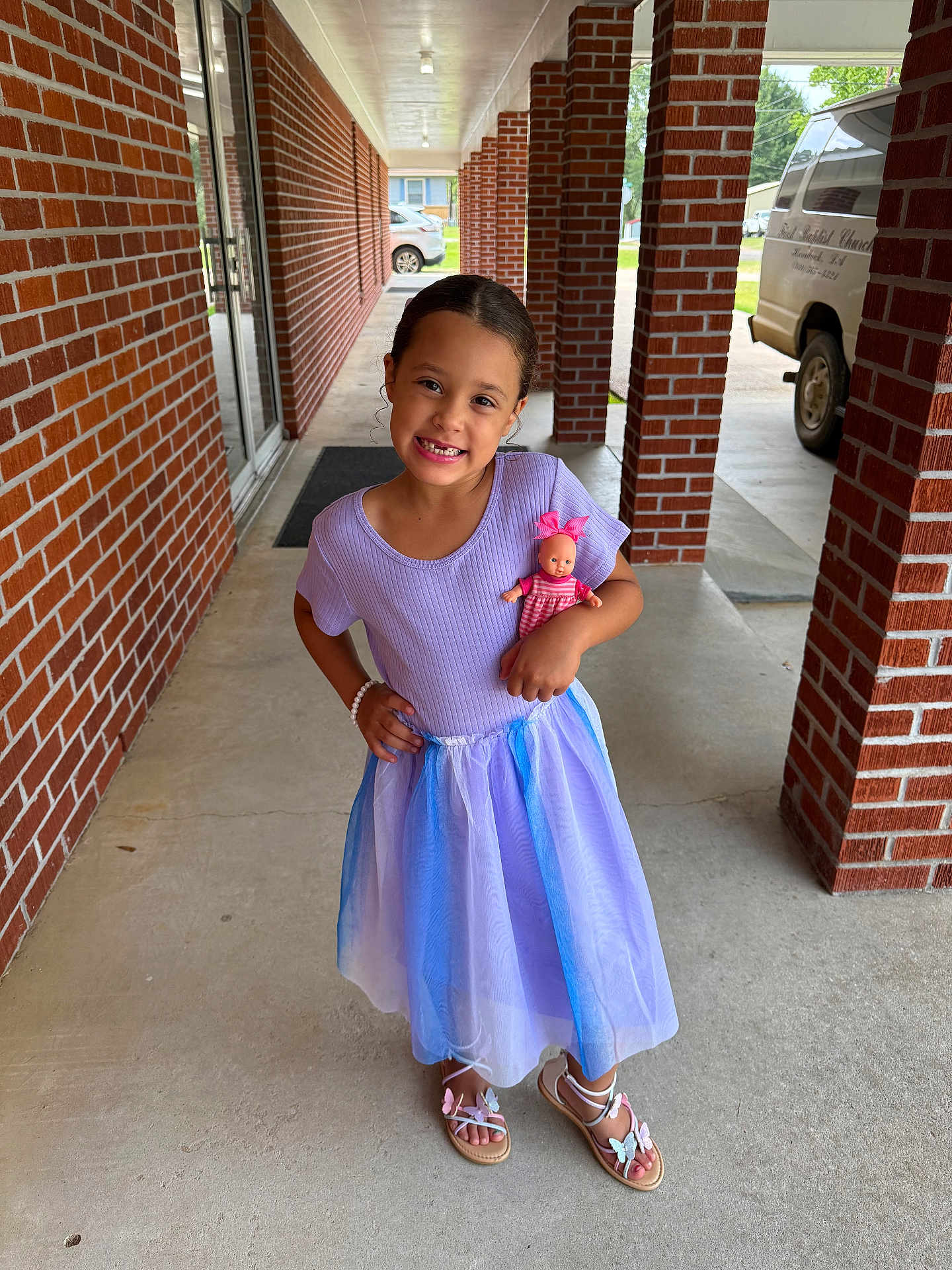 Raelynn is registered to the contest to win money with this photo: child, girl, smiling, dress, doll, sandals, bracelet, brick_wall, sidewalk, outdoor, portrait, happy, person, toy, footwear, building, window, door, concrete, sunlight
