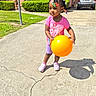 toddler, child, girl, orange_ball, pink_shirt, purple_shorts, colorful_hair_beads, outdoor, driveway, residential_house, green_hedge, white_suv, sunny, shadow, concrete, smiling, playing, casual_clothing, daytime, person