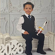 Austin joined the competition — help win amazing prizes! child, boy, graduation, diploma, suit, tie, smile, bench, numbers, 2026, portrait, formal, indoor, curly_hair, white_shirt, black_pants, black_shoes, decorative_wall, celebration, happy