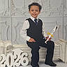 child, boy, graduation, diploma, suit, tie, smile, bench, numbers, 2026, portrait, formal, indoor, curly_hair, white_shirt, black_pants, black_shoes, decorative_wall, celebration, happy