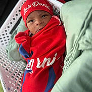 Luis joined the competition — help win amazing prizes! baby, infant, red_blanket, red_hat, basket, white_basket, face, expression, wrapped, soft_fabric, green_fabric, newborn, small, cute, indoors, portrait, clothing, lying_down, young_child, headwear