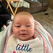 Aiyana is registered to the contest to win money with this photo: baby, infant, bib, blanket, face, smile, indoor, living_room, couch, table, wooden_table, child, person, clothing, cute, sleepy, soft, home, comfort, portrait