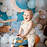 baby, child, toy_airplane, wooden_toy, overalls, balloons, crescent_moon, teddy_bear, smiling, indoor, floor, blue_background, stars, clouds, playful, cute, portrait, happy, flooring, decor