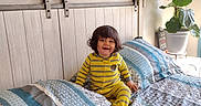 Aziel joined the competition — help win amazing prizes! toddler, child, bed, bedding, pajamas, yellow_stripes, headboard, plant, pillow, blanket, indoor, smiling, happy, hair, furniture, home, light, window, wall_art, room