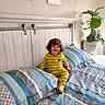 toddler, child, bed, bedding, pajamas, yellow_stripes, headboard, plant, pillow, blanket, indoor, smiling, happy, hair, furniture, home, light, window, wall_art, room