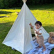 Daniel is registered to the contest to win money with this photo: child, toddler, outdoor, teepee, tent, grass, garden, portrait, standing, smiling, teddy_bear, blanket, playhouse, nature, wooden_poles, sunlight, summer, clothing, wilderness, camping