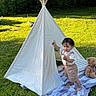child, toddler, outdoor, teepee, tent, grass, garden, portrait, standing, smiling, teddy_bear, blanket, playhouse, nature, wooden_poles, sunlight, summer, clothing, wilderness, camping