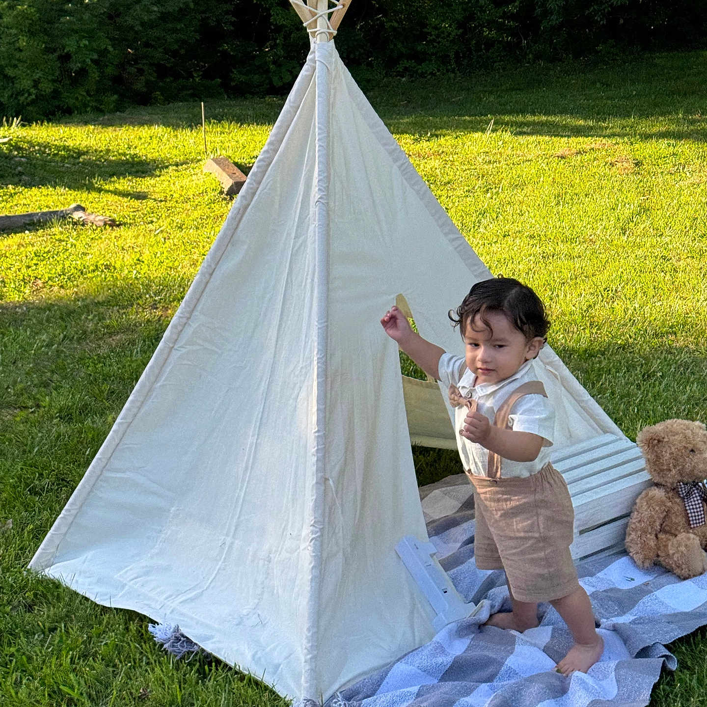 Daniel is registered to the contest to win money with this photo: child, toddler, outdoor, teepee, tent, grass, garden, portrait, standing, smiling, teddy_bear, blanket, playhouse, nature, wooden_poles, sunlight, summer, clothing, wilderness, camping
