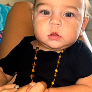 Inaya participe au concours pour gagner de l'argent avec cette photo : baby, child, infant, face, hand, holding, black_clothing, necklace, beads, skin, eyes, person, portrait, closeup, cute, indoor, human, expression, young, chubby_cheeks