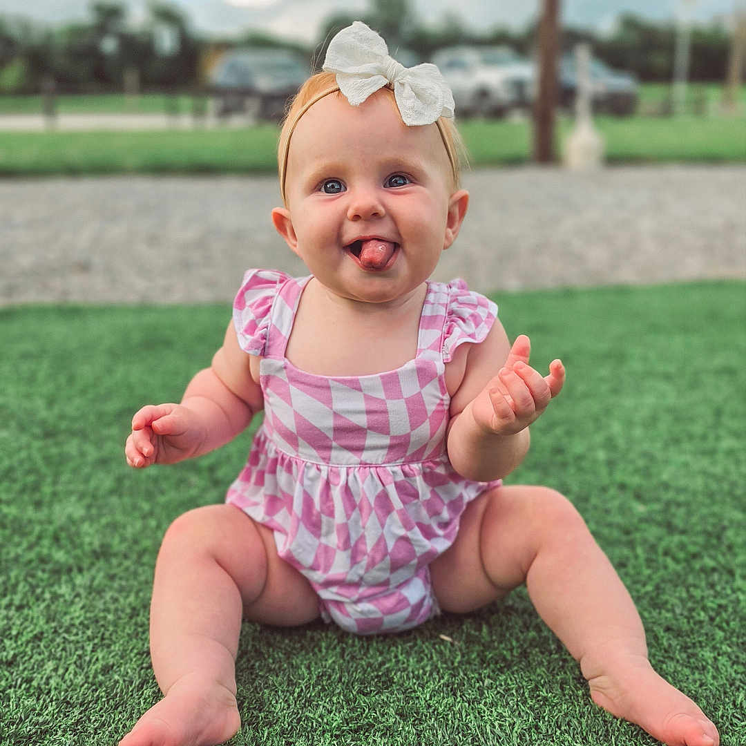 Bexley is registered to the contest to win money with this photo: baby, child, girl, tongue_out, bow, headband, pink_clothing, checkered_pattern, grass, outdoor, nature, happy, playful, sitting, cute, infant, portrait, daylight, fun, smile