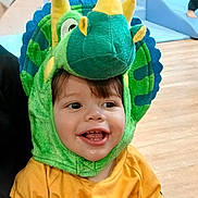 Othman a rejoint le concours — aidez-le/la à gagner de superbes lots ! child, toddler, smile, dinosaur_costume, green, yellow, hood, plush, cute, happy, eyes, teeth, tongue, indoor, wooden_floor, playroom, clothing, portrait, sitting, hand