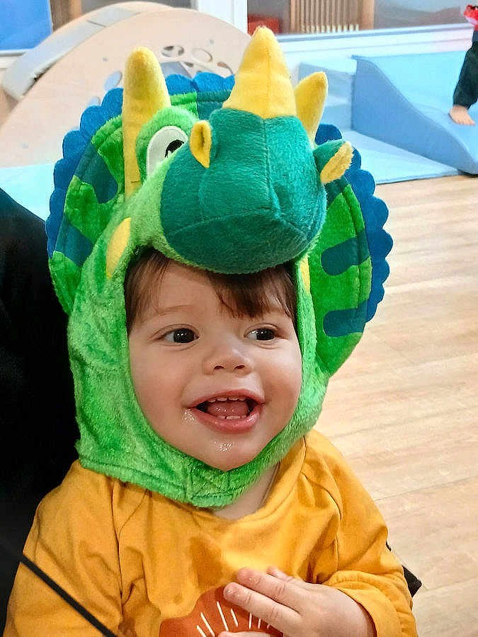 Othman a rejoint le concours — aidez-le/la à gagner de superbes lots ! child, toddler, smile, dinosaur_costume, green, yellow, hood, plush, cute, happy, eyes, teeth, tongue, indoor, wooden_floor, playroom, clothing, portrait, sitting, hand