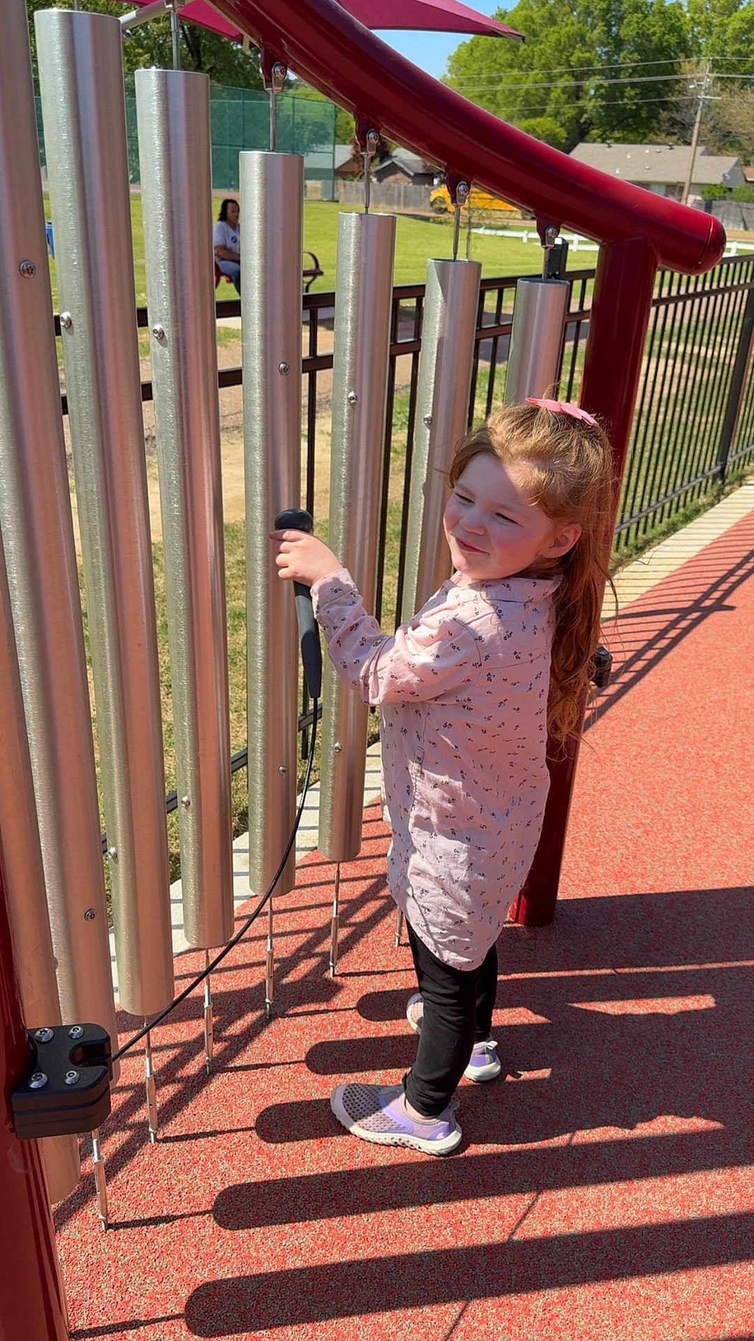 Kailynn is registered to the contest to win money with this photo: child, girl, outdoor, musical_instrument, xylophone, playground, music, toy, red_ground, shadows, sunlight, park, fence, long_hair, pink_bow, casual_clothing, black_pants, white_shoes, happy, playing