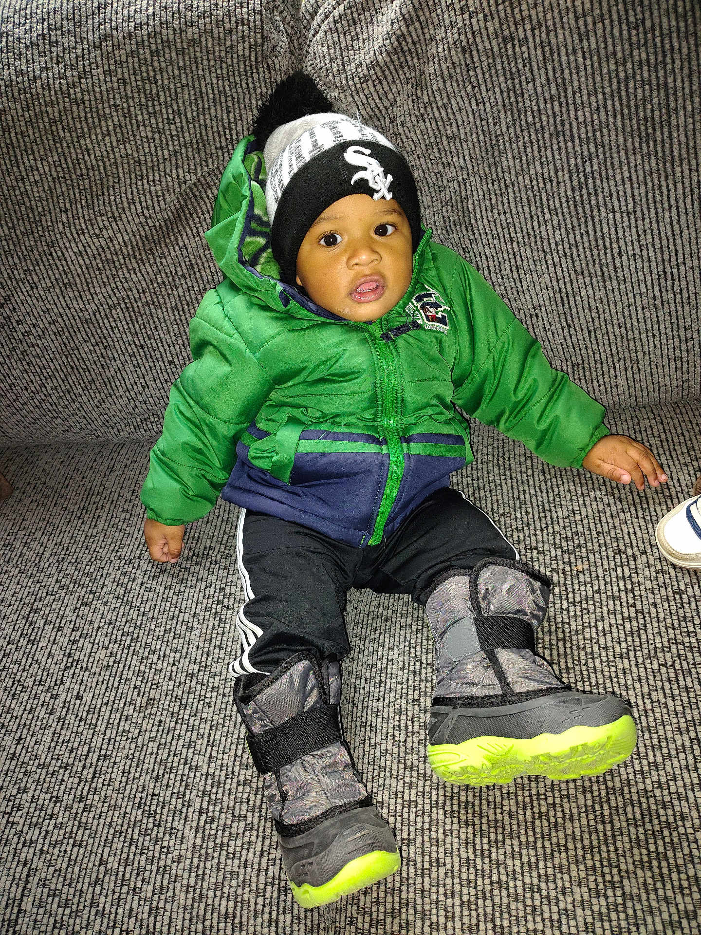 Tayzon joined the competition — help win amazing prizes! toddler, child, green_jacket, winter_hat, boots, couch, indoor, portrait, sitting, curious, warm_clothing, footwear, black_pants, textile, face, person, young_child, winter_wear, cozy, home