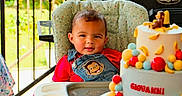 Giovanni is registered to the contest to win money with this photo: toddler, child, high_chair, birthday_cake, cake, colorful, outdoor, grill, party, celebration, denim_overall, monkey_patch, food, table, greenery, sunlight, baby, face, smile, curious